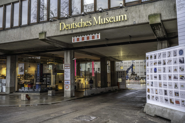 Außenansicht des Deutschen Museums in Berlin: großes Glasgebäude mit Säulen, Namensschild, Informationsschild und umgebende Strukturen, Bäume und Himmel.
