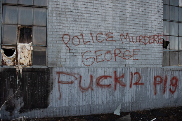 Ein Gebäude mit Graffiti, das "Polizei Mord George Fuck 12 P19" an der Seite trägt und Fenster sowie verschiedene Gegenstände vorne zeigt.