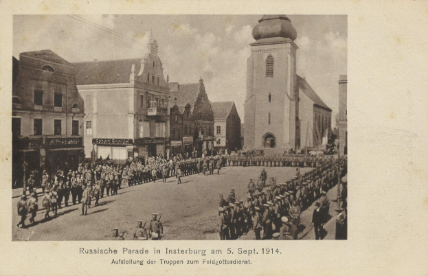 Ein Schwarz-Weiß-Foto eines Umzugs in Insterburg am 5. September 1914, das viele Menschen, Gebäude mit Fenstern im Hintergrund und bewölkten Himmel zeigt, mit Text am unteren Bildrand.