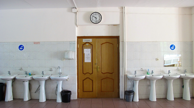 Badezimmer mit einer Reihe von Waschbecken mit Wasserhähnen, Bodenmülltonnen, einer Tür im Hintergrund, einer Wanduhr und einem Spiegel auf der rechten Seite.