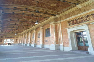 Langer Flur mit gefliestem Boden, Säulen, Türen, Wandrahmen, Deckenleuchten und mehreren Menschen, der einem belebten Rathausinneren ähnelt.