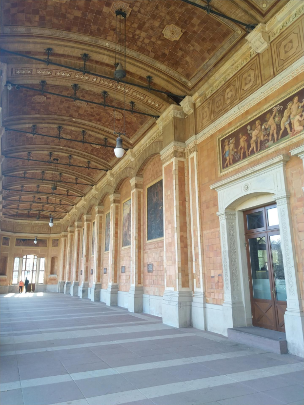 Langer Flur mit gefliestem Boden, Säulen, Türen, Wandrahmen, Deckenleuchten und mehreren Menschen, der einem belebten Rathausinneren ähnelt.