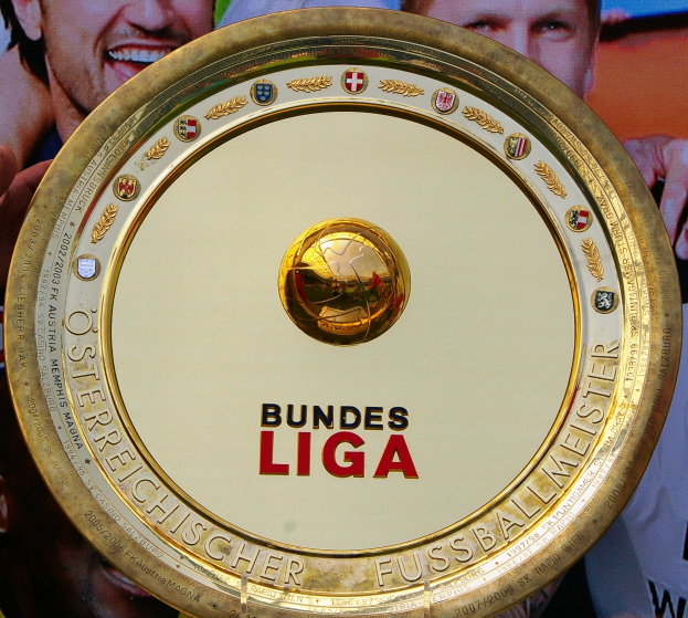Ein Mann hält einen goldenen Schild mit der Aufschrift "Bundesliga", im Hintergrund eine Fahne mit drei Personen.