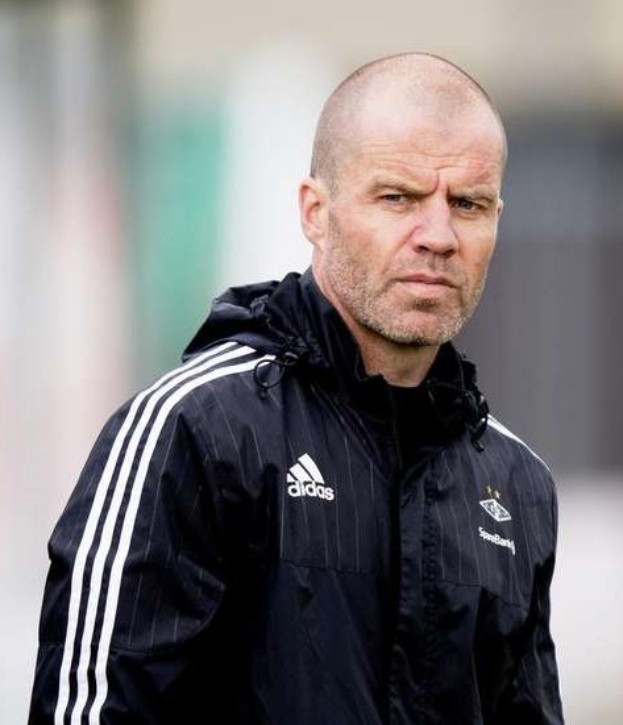 Ein kahlköpfiger Mann in einer schwarzen Jacke steht auf einem Fussballfeld und trägt eine marineblaue Jacke mit weißen Streifen und einem weißen Kragen. Seine Hände sind verschränkt und er sieht ernst aus.