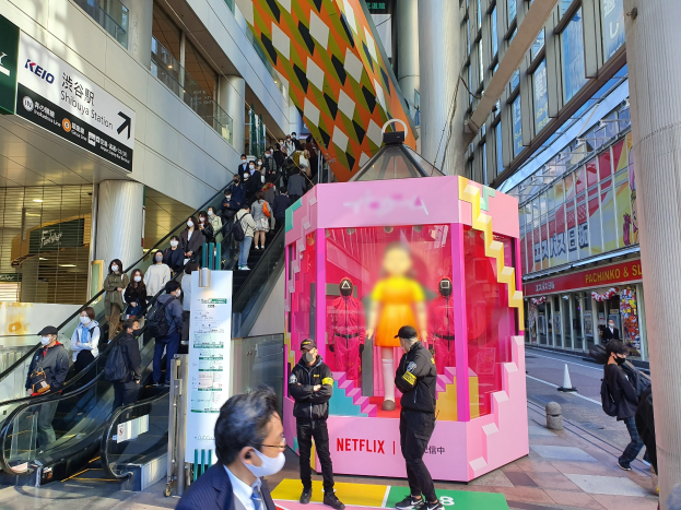 Eine Gruppe von Menschen steht vor einem Gebäude mit Glaswänden und Texttafeln, einige tragen Masken, während eine Rolltreppe mit Menschen daneben fährt; eine pinkfarbene Box in der Mitte zeigt Schaufensterpuppen und trägt den Text "Netflix's neuer Netflix-Schalter in Tokyo."