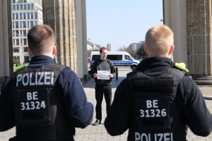 Drei Polizeibeamte stehen vor einem Gebäude, wobei ein Mann in der Mitte ein Papier hält, flankiert von Säulen, mit Fahrzeugen, Menschen, Bäumen und Gebäuden im Hintergrund.