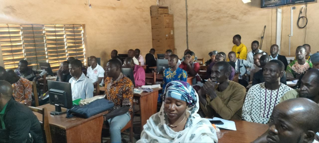 Eine Gruppe von Menschen sitzt an Tischen in einem Klassenzimmer mit Monitoren, Taschen, Büchern und Papieren und nimmt an einem Techniktraining teil; links ist ein Fenster, und Kartons und ein Fernseher sind an der Wand im Hintergrund zu sehen.