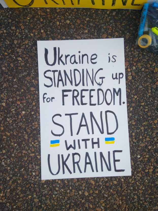Weißer Papierbogen mit dem Text "Ukraine steht für Freiheit, steh mit Ukraine" auf dem Boden vor einer gelben Fahne, mit einer Plastikabdeckung mit verschiedenen Gegenständen rechts.