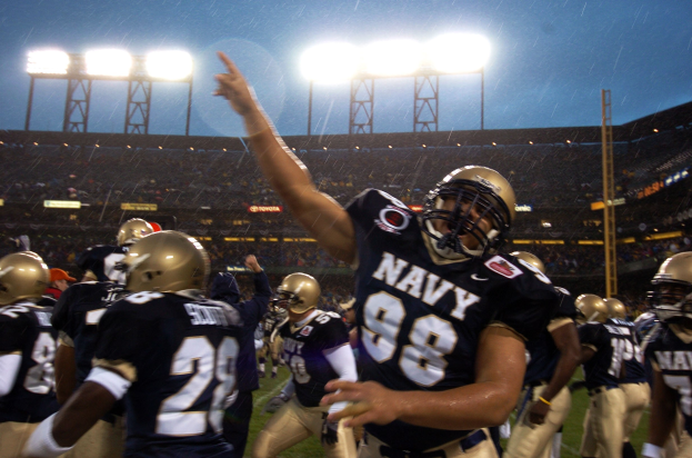 Navy-Fußballspieler in marineblauen und weißen Uniformen feiern ein Touchdown auf einem regennassen Rasenfeld, mit Zuschauern und Stadionbeleuchtung im Hintergrund.