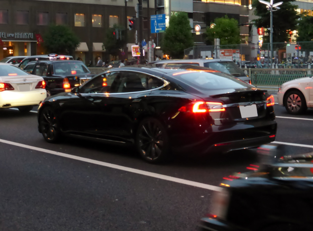 Schwarzer Tesla Model S fährt nachts auf einer Stadtstraße mit anderen Fahrzeugen, Gebäuden, Bäumen und Fußgängern im Hintergrund.