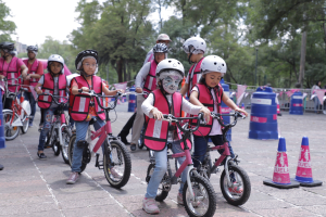 Kinder mit Helmen fahren auf einer mit Verkehrskegeln markierten Straße Fahrräder, einige mit Gesichtsbemalung, vor einem Hintergrund aus Bäumen und einem klaren Himmel.