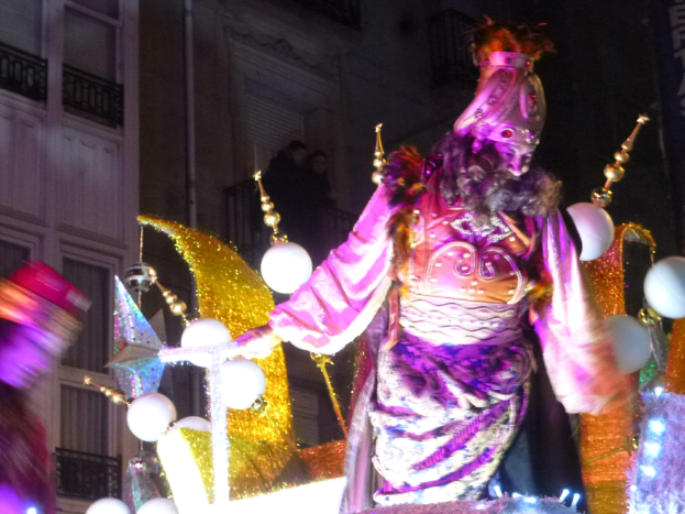 Ein Mann in einem Kostüm steht auf einem dekorierten, beleuchteten Festumzugswagen in einer Stadtparade, mit einem Gebäude mit Fenstern und Geländern im Hintergrund.