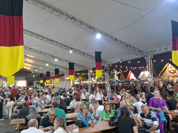 Eine große Gruppe von Menschen sitzt an Tischen unter einem Zelt auf dem Oktoberfest in München, Deutschland, mit einigen stehenden in der Nähe und festlichen Décor im Hintergrund.