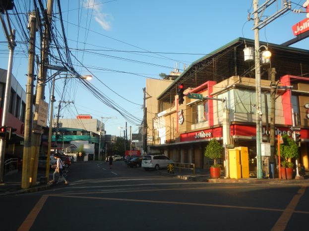 Eine belebte Stadtstraße mit Autos, Fußgängern, Strommasten, Ampeln, Gebäuden mit Schildern, Topfpflanzen und einer bewölkten Himmel.