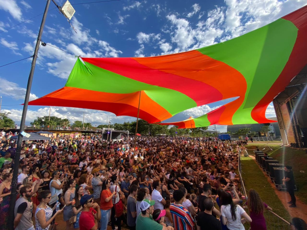 Eine große Menge steht vor einer Regenbogenflagge mit einem Gebäude, Lautsprechern auf der Bühne, Bäumen und Wolken im Hintergrund bei der 10-jährigen Feier eines Festivals.