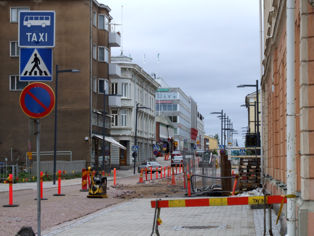 Stadtstraße mit Gebäuden, Verkehrsschildern, Fahrzeugen, Bäumen und einer Baustelle im Hintergrund unter einem bewölkten Himmel.