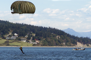 Eine Person gleitet mit einem Gleitschirm über ein Gewässer, mit einem Boot auf der rechten Seite, Bäume, Gebäude, Hügel und einen klaren blauen Himmel im Hintergrund.