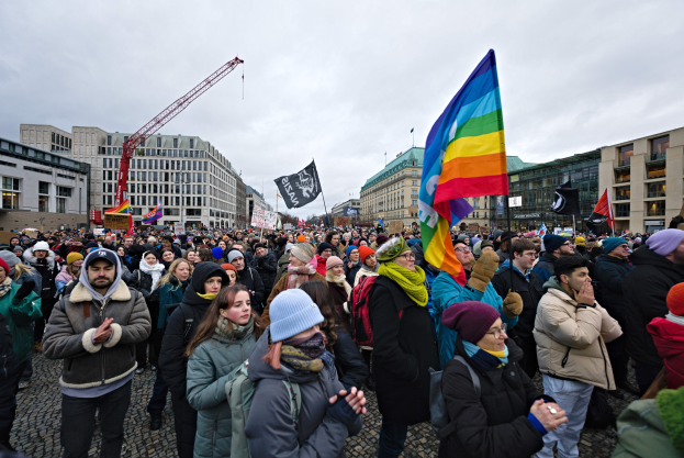 Große Gruppe von Menschen mit Fahnen und Spruchbändern, darunter "Lgbtq+ Rechte Demonstration in Berlin", vor einem Gebäude mit Fenstern, einem Kran und einem bewölkten Himmel.