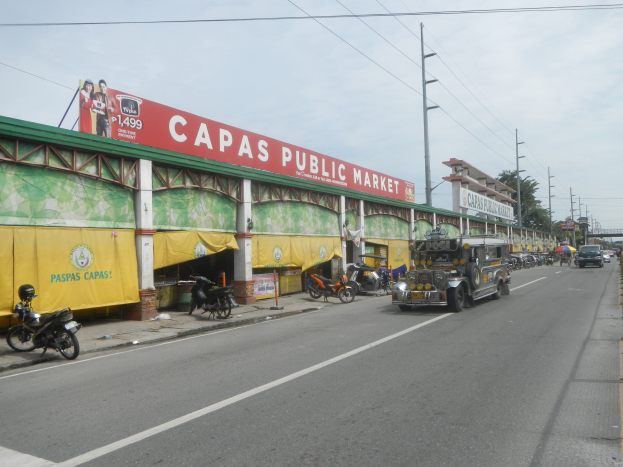 Eine belebte Stadtstraße mit Fahrzeugen, einem Gehweg, Strommasten, Gebäuden, Bäumen, einer bewölkten Himmel und einem Gebäude im Vordergrund mit der Aufschrift "Capas Public Market".
