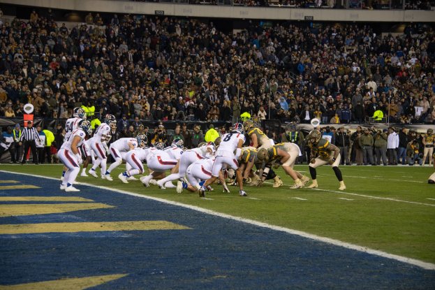 Ein Fußballspiel im Gange mit Spielern auf dem Boden in Sportbekleidung und Helmen, aus der Perspektive einer Tribüne mit Zuschauern, Geländern und Beschriftungen im Hintergrund.