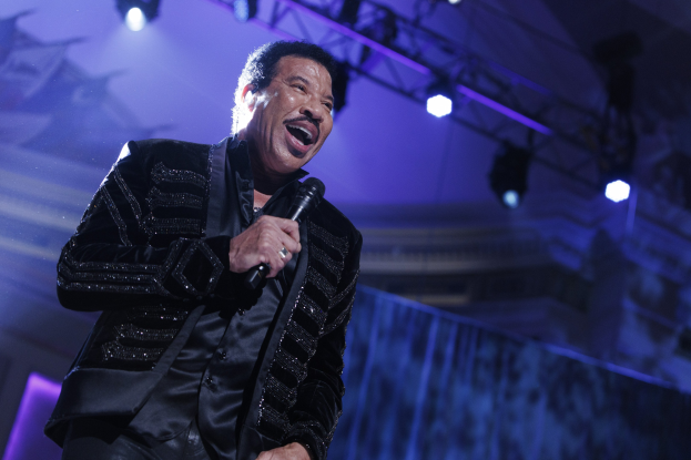 Lionel Richie performt bei den Grammys in einem schwarzen Kleid und hält ein Mikrofon auf einer hell erleuchteten Bühne.
