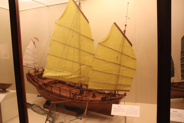 Ein hölzernes Modellsegelboot in einer Glasvitrine mit informativen Tafeln im Hintergrund.