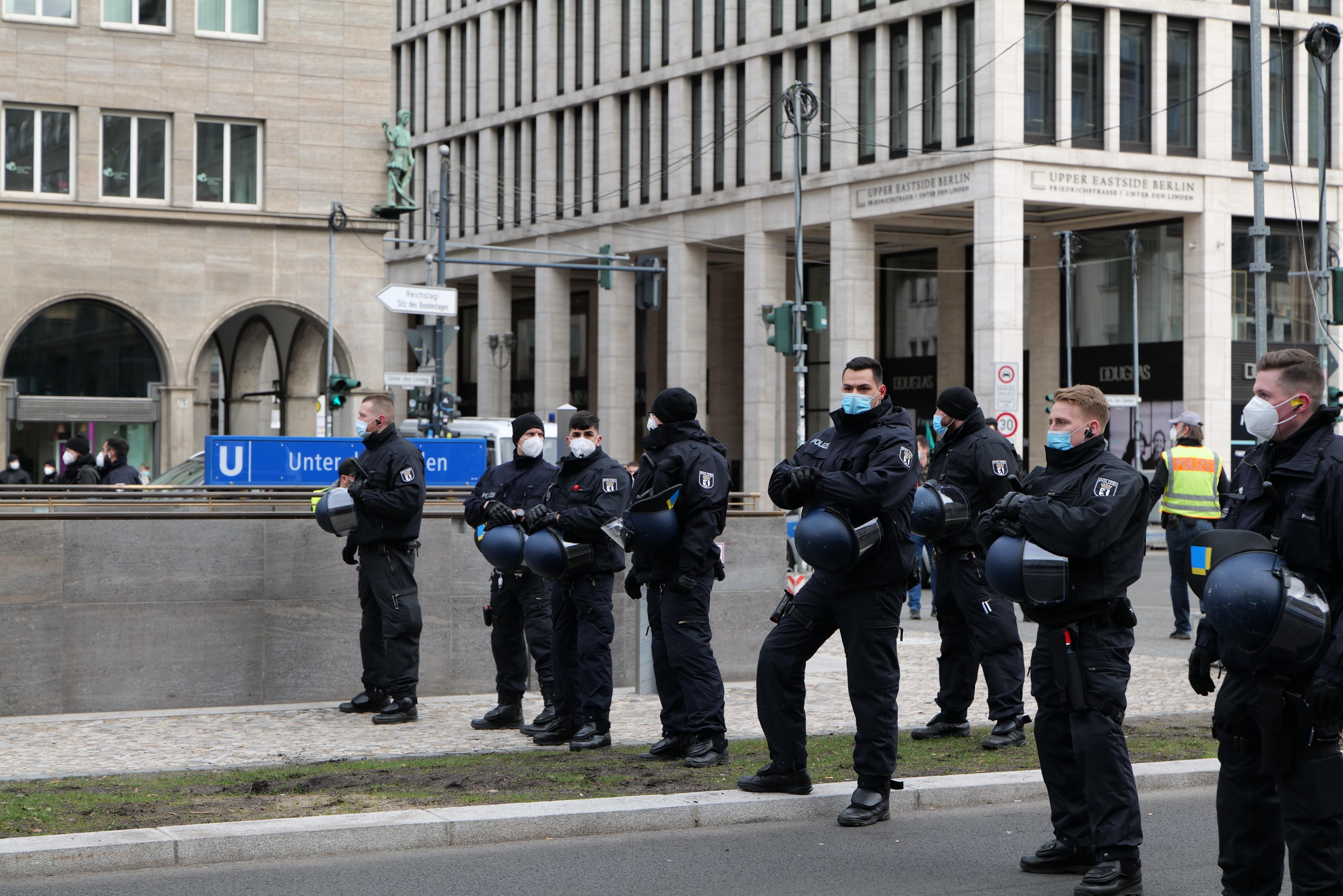 Polizeibeamte in schwarzen Uniformen und Masken stehen vor einem Gebäude mit Glasfenstern und Säulen in Berlin, Deutschland, einige halten Helme, mit Laternen, Verkehrsampeln, Schildern mit Text und einer Statue im Hintergrund.