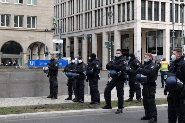 Polizeibeamte in schwarzen Uniformen und Masken stehen vor einem Gebäude mit Glasfenstern und Säulen in Berlin, Deutschland, einige halten Helme, mit Laternen, Verkehrsampeln, Schildern mit Text und einer Statue im Hintergrund.