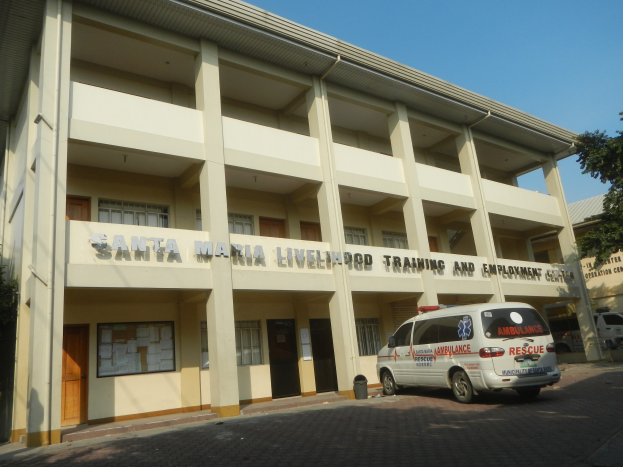 Außenansicht des Santa Maria Lebensmittel-Trainings- und Beschäftigungszentrums mit Fenstern, Türen und Beschriftung, umgeben von Bäumen und Fahrzeugen auf einer Straße unter einem klaren blauen Himmel.