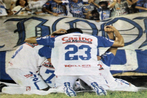 Zwei Fußballspieler feiern ein Tor auf einem Feld, umgeben von einer Gruppe von Menschen, mit einer Person, die eine Kamera hält, einem Banner mit Text, Stühlen und einer Wand im Hintergrund.