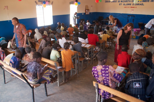 Kinder auf Bänken im Vordergrund, mit Erwachsenen, die sie führen, und einem Hintergrund mit Stühlen, einer Tür, einem Fenster, einem Tisch und Text oben.