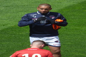 Ein Mann in einem Sportoutfit steht auf einem gr├╝nen Feld und h├Ąlt eine Flasche in einer Hand und ein oranges Tuch in der anderen.