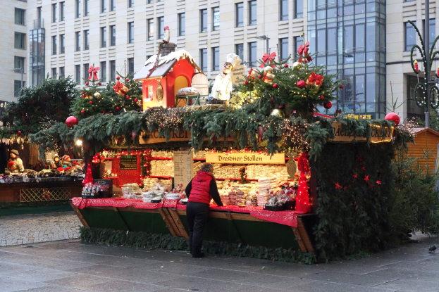Eine Person steht vor einem Weihnachtsmarktstand auf einem Stadtplatz, umgeben von festlichen Dekorationen und anderen Menschen, mit Gebäuden und einem Laternenmast im Hintergrund.