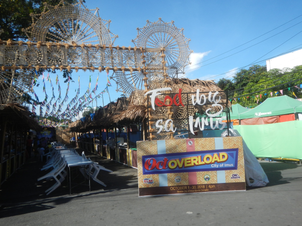 Eine Karnevalszene mit einem großen Riesenrad in der Mitte, umgeben von Hütten, Tischen, Stühlen, einem Schild, Dekorationen, Bäumen, Überlandleitungen und einem bewölkten Himmel.