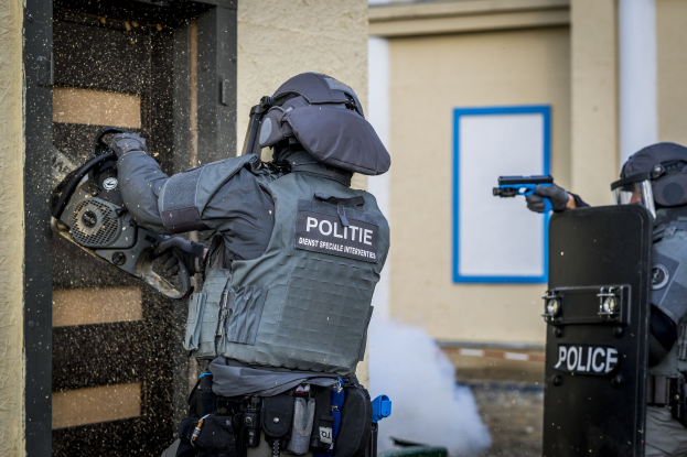 Zwei Polizisten in Einsatzkleidung stehen vor einem Gebäude, einer hält eine Waffe und der andere ein Objekt, mit einer Tür links und einer Tafel mit Text an der Wand dahinter.