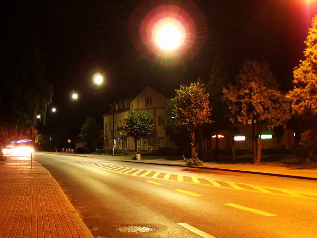 Eine nächtliche Stadtstraße mit beleuchteten Straßenlaternen, ein Gehweg auf der linken Seite, Bäume und Gebäude im Hintergrund und ein dunkler Himmel darüber.