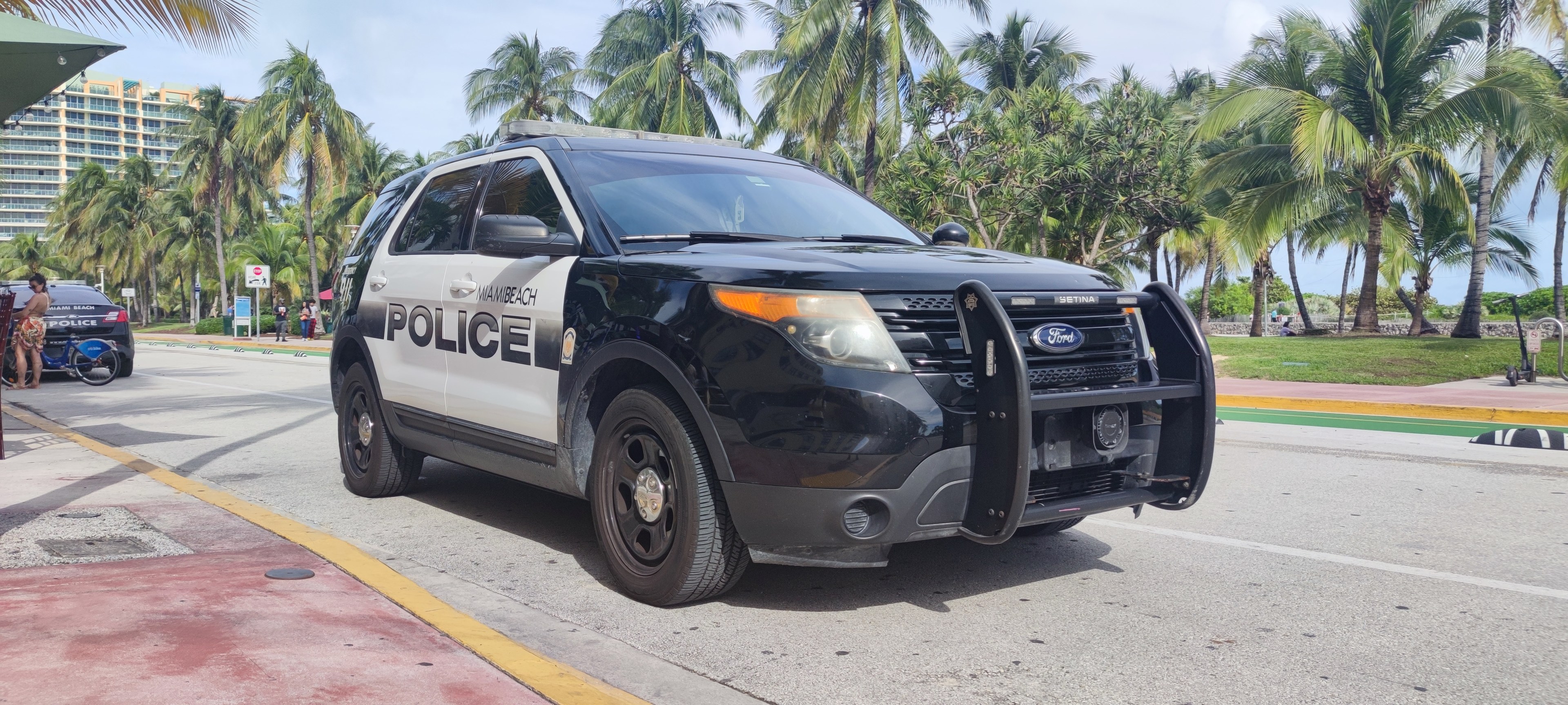 Ein Polizeiwagen am Straßenrand geparkt, umgeben von Menschen, Fahrrädern, Gras, Bäumen, Gebäuden und einem klaren blauen Himmel, mit seinem Vorderrad sichtbar.