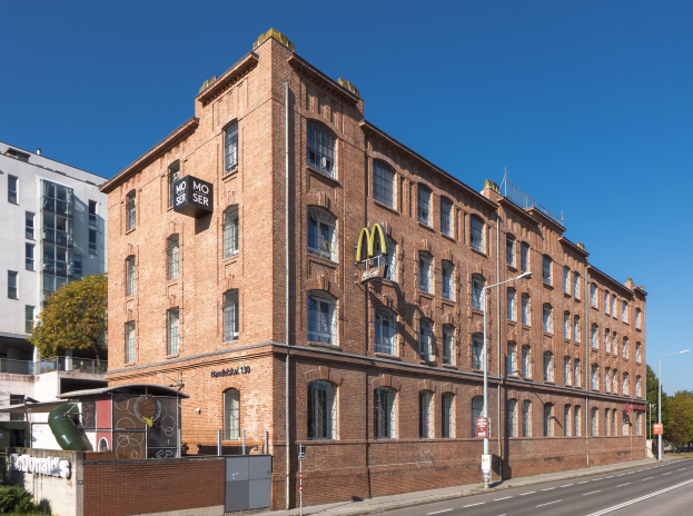 Ein großes McDonald's-Restaurantgebäude aus Backstein an einer Straßenecke in Berlin, Deutschland, mit umliegender Straßeninfrastruktur, Fahrzeugen, Grünflächen und einem bewölkten Himmel.
