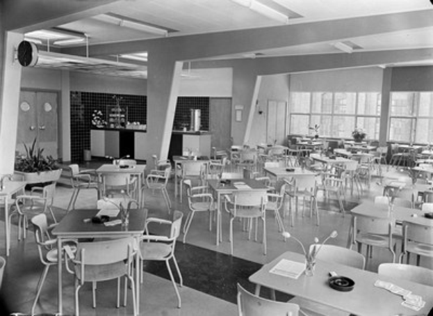 Schwarz-weiß-Foto einer Cafeteria mit Tischen und Stühlen, Tellern, Besteck, Gläsern und Blumenvasen auf den Tischen und einer Wand mit Türen, Fenstern und Deckenleuchten im Hintergrund.