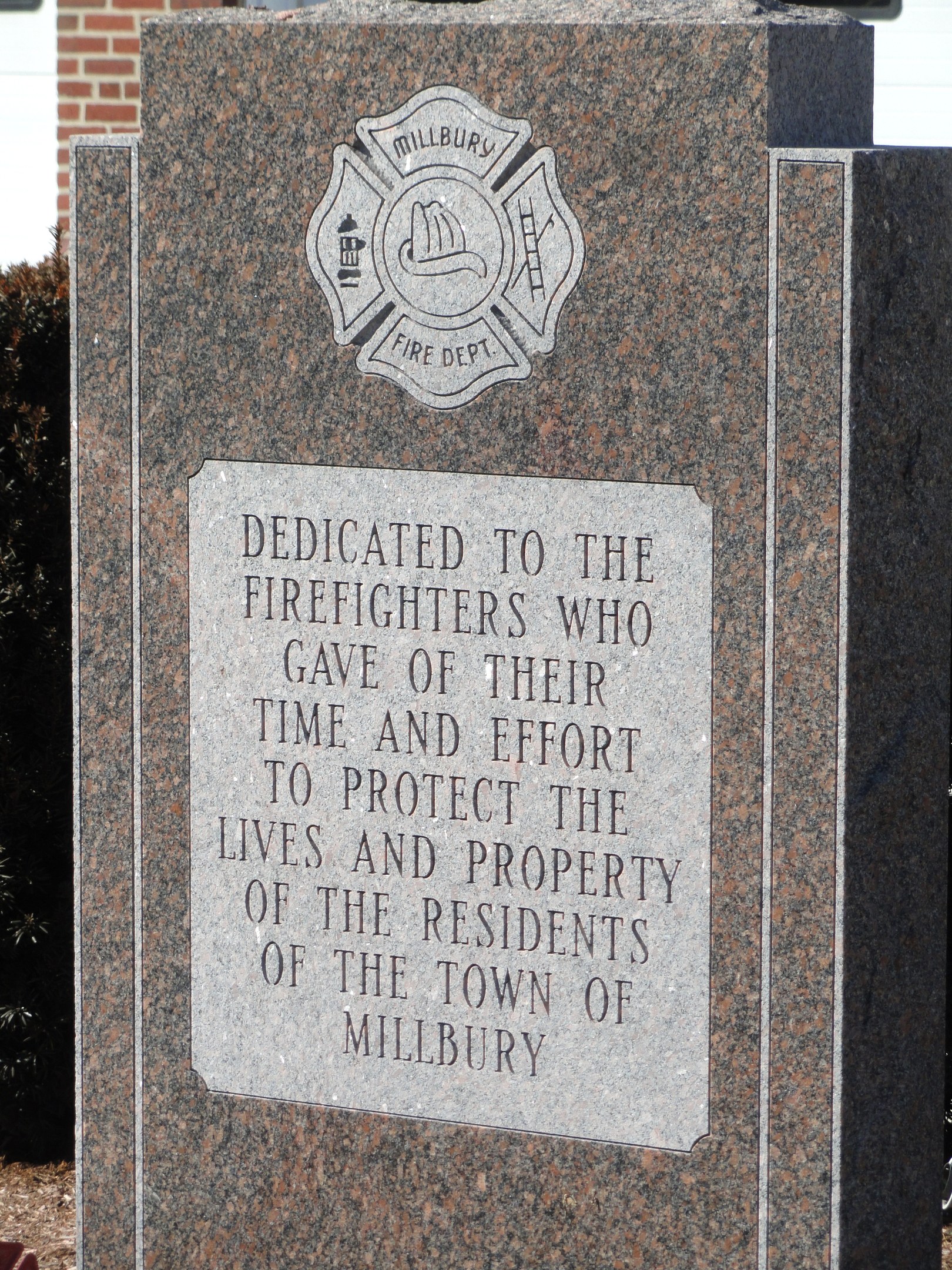 Denkmalstein mit einer Gedenktafel für Feuerwehrleute in Millbury, vor einem Backsteingebäude inmitten von Grünanlagen.