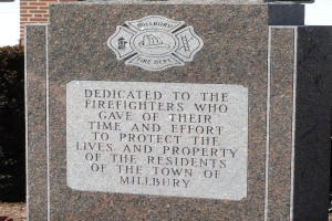 Denkmalstein mit einer Gedenktafel für Feuerwehrleute in Millbury, vor einem Backsteingebäude inmitten von Grünanlagen.