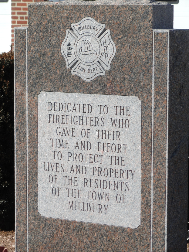 Denkmalstein mit einer Gedenktafel für Feuerwehrleute in Millbury, vor einem Backsteingebäude inmitten von Grünanlagen.