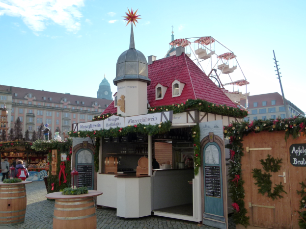 Ein kleines Gebäude mit einem roten Dach und einem Riesenrad davor, umgeben von Festdekorationen und Menschen bei einem Outdoor-Event.
