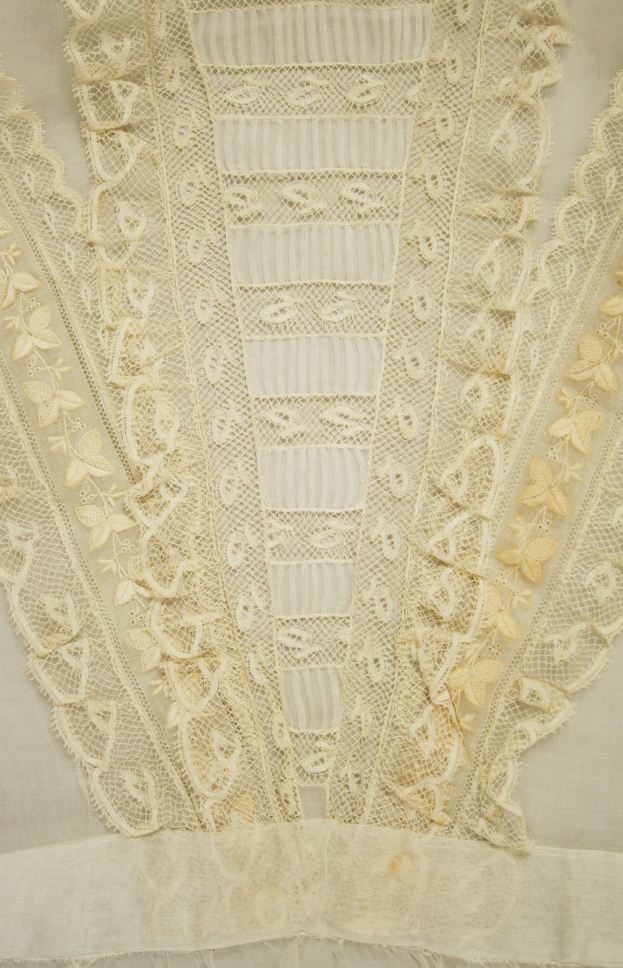 Nahaufnahme eines weißen Kleides mit zarter, detaillierter Spitzenverzierung auf einem leichten, luftigen Stoff.