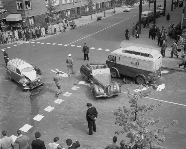 Eine Schwarz-Weiß-Szene eines Autounfalls an einer Straßenecke, mit mehreren Fahrzeugen, darunter ein Van, einer Gruppe von Menschen drumherum und Gebäuden, Bäumen, Masten und Texttafeln im Hintergrund.