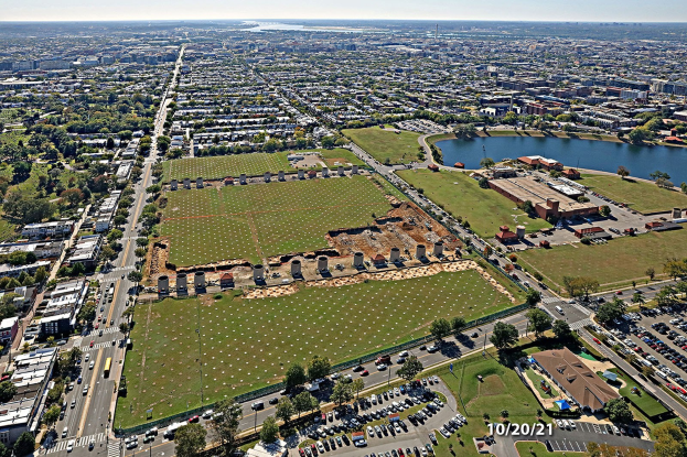 Luftaufnahme einer Stadt mit Gebäuden, Bäumen, Gras, Straßen mit Fahrzeugen, Wasser und Wolken, die einen großen zentralen Platz als Parkplatz zeigt.