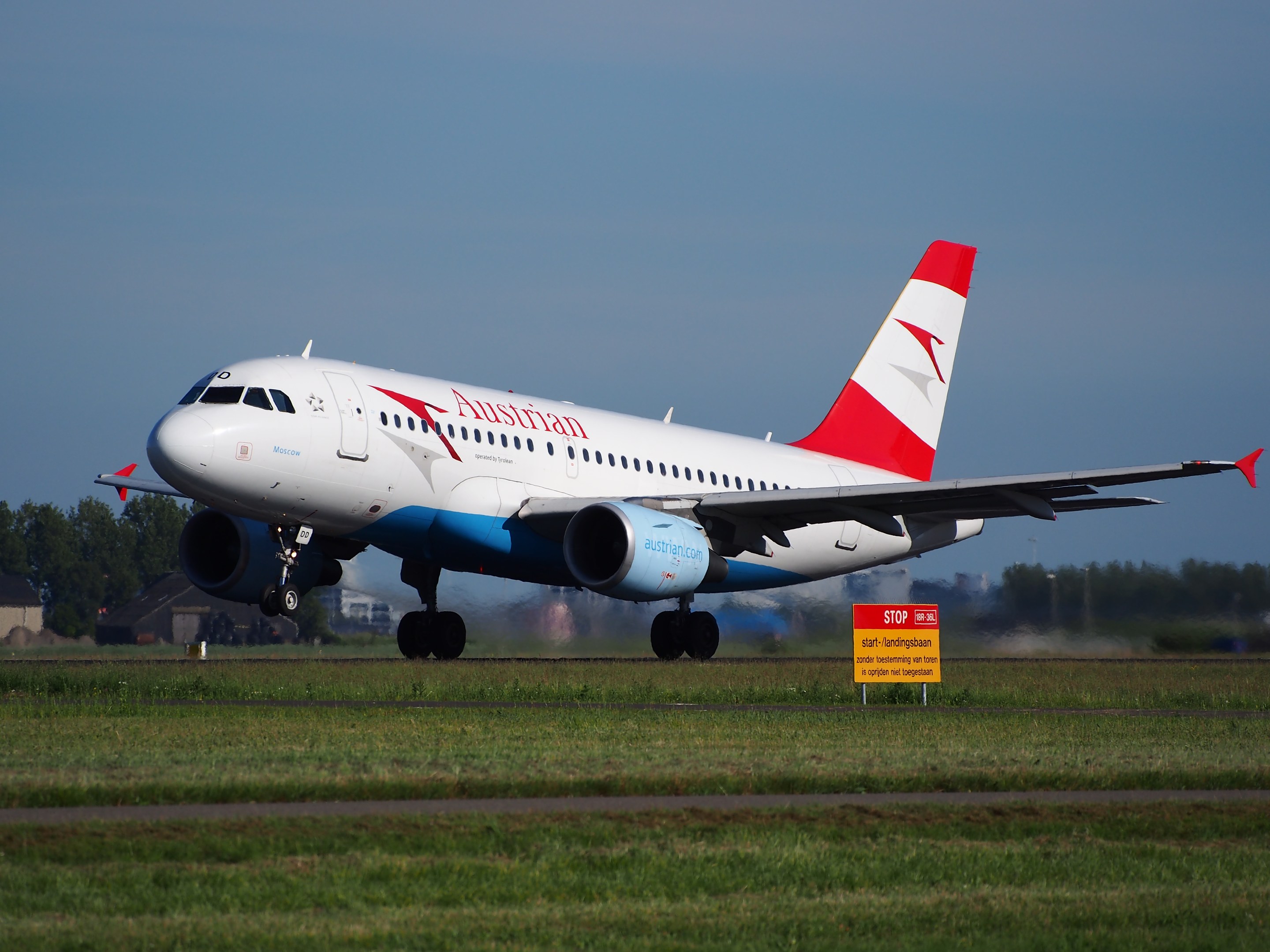 Ein Airbus A320-200 der Austrian Airlines startet vom Frankfurter Flughafen mit einer Tafel im Vordergrund, Gras, Bäumen, Gebäuden und einem klaren blauen Himmel.