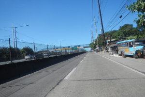 Arterielle Straße mit Fahrzeugen, Strommasten, einem Zaun, Bäumen, Straßenlaternen, einer Brücke im Hintergrund, einem blauen Himmel und einem geparkten Bus.