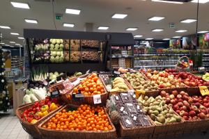 Ein Geschäft mit Körben voller Obst und Gemüse, jeder mit Preisschild, einem geschmückten Weihnachtsbaum links und Regalen mit anderen Artikeln im Hintergrund.