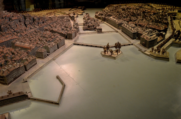 Ein detailliertes Stadtmodell in einem Museum ausgestellt, mit Gebäuden, Bäumen und anderen Objekten, umgeben von Menschen, die es betrachten.